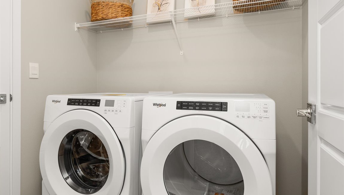 A laundry room with a washer and dryer under a shelf holding up several art pieces and baskets.