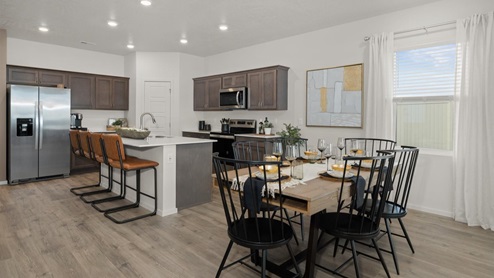 A dining area with a wooden table and black chairs in the foreground and a kitchen with brown wooden cabinets, a large island with three stools, a pantry, and a refridgerator to the left. There is abstract art on the right wall next to a window letting in natural light framed by sheer white curtains.