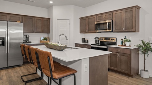 A kitchen with brown wooden cabinets, a refridgerator to the left, a pantry, and a kitchen island in the middle with three burnt orange stools.
