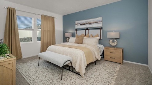 A bedroom with a blue accent wall featuring a bed with beige sheets, abstract art above the headboard, two nightstands with lamps, a dresser against the left wall, a large ornamental rug, and two large windows letting in natural light framed by tan curtains.