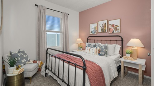 A bedroom with a pink accent wall featuring a bed with white and pink sheets and a metal bedframe. There are two side tables with lamps and art on the wall above the headboard. There's an accent chair in the left corner and a window letting in natural light framed by sheer grey curtains.