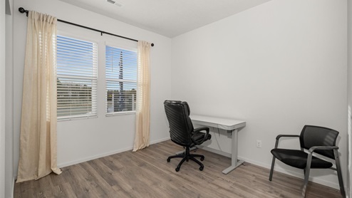 A room with a white office desk and black office chair, a black chair in the right corner, and two large windows letting in natural light framed by beige curtains.