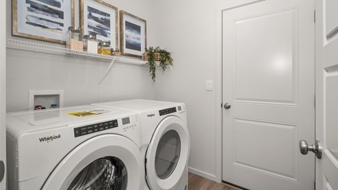 A laundry room with a washer and dryer under a shelf holding up several art pieces and a plant.