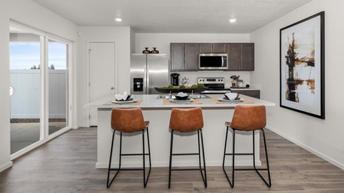 A kitchen with brown wood cabinets and stainless steel appliances, there's a large island in the middle with three burnt orange stools, a large picture frame on the right wall, and to the left there's glass sliding doors leading to the backyard.