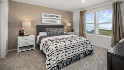 A bedroom with a bed with white patterned sheets in a black bedframe, there are two nightstands with lamps, and two large windows letting in natural light framed by dark beige curtains.
