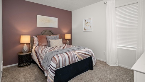 A bedroom with a bed with burgundy patterned sheets and two nightstands with lamps on them, there's a burgundy accent wall and multiple picture frames.