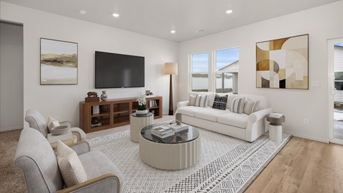 A living area with a large white patterned rug and white couch and grey accent chairs. There's a white coffee table in the middle and a television mounted on the left wall. There's abstract art hanging on the walls and two windows letting in natural light.
