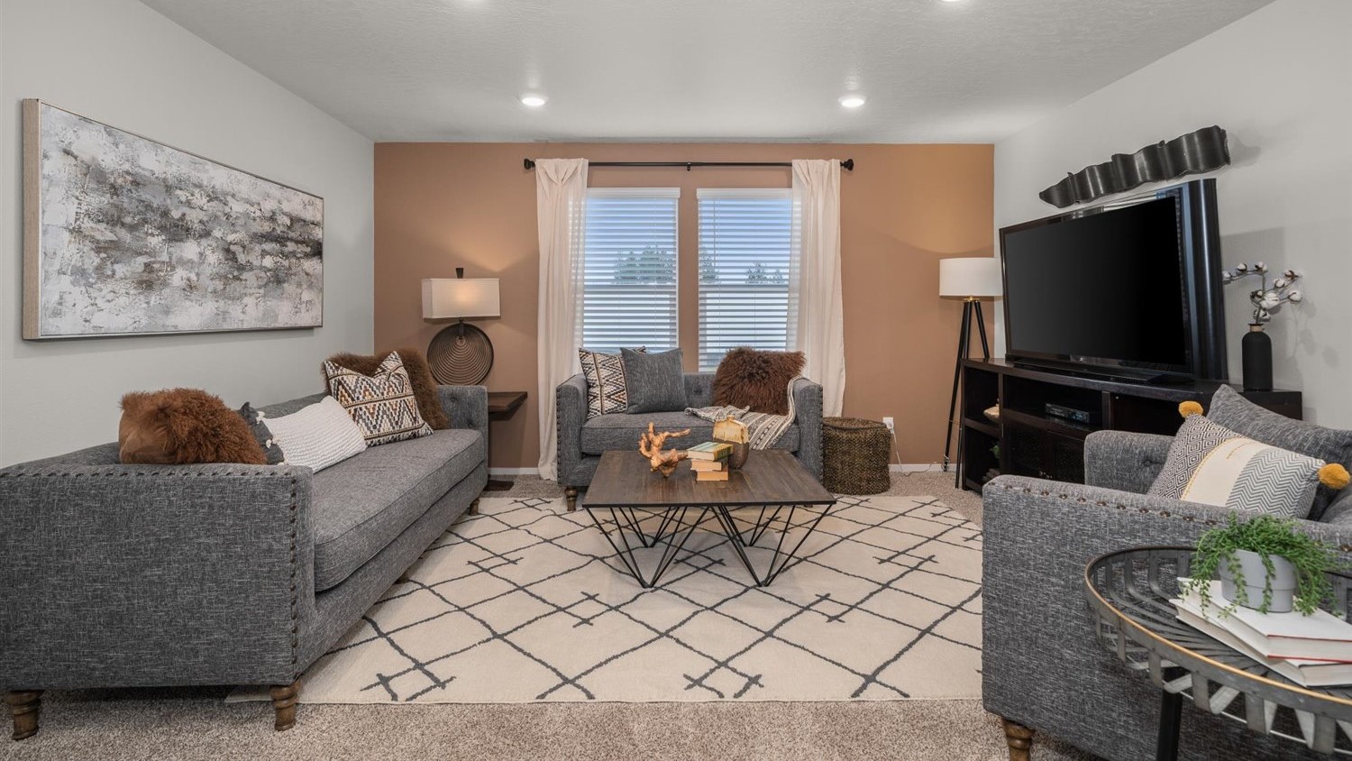 A living area with a large off white patterned rug and grey couch and accent chairs, there's a tan accent wall with two large windows letting in natural light framed by beige curtains. There is abstract art on the left wall and a large television on the right wall.