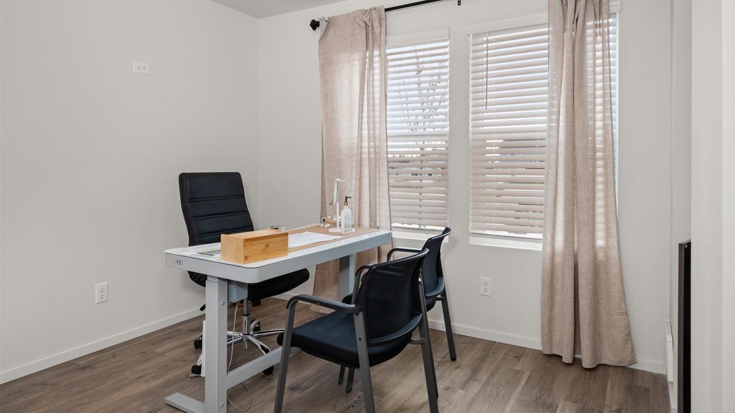 A room with a white office desk and a black chair, there are two black accent chairs, and two large windows with blinds covering them accented by sheer beige curtains.