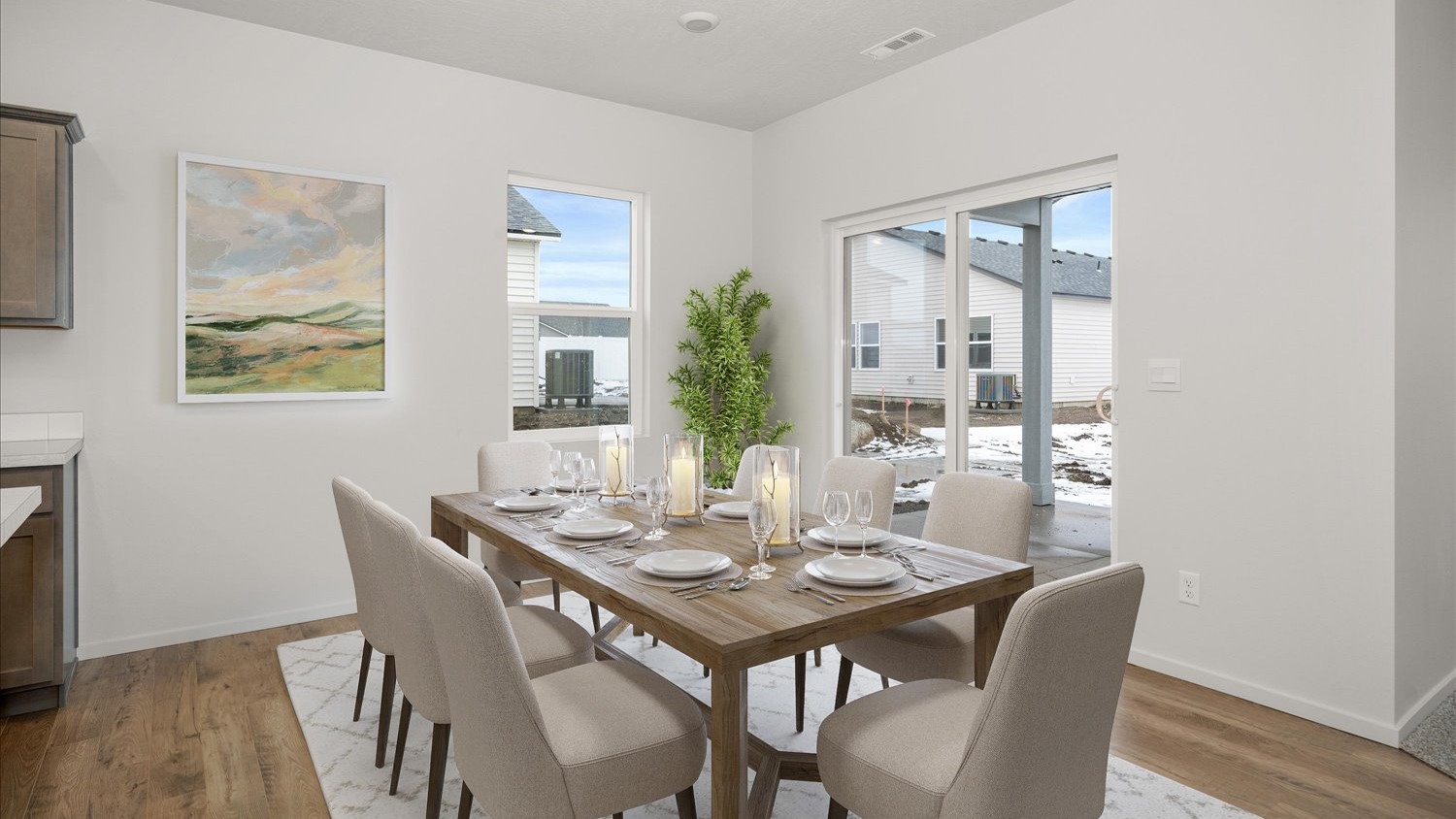 A dining area with a wooden dining table, beige chairs, art hanging on the wall to the left next to a window letting in natural light, and glass sliding doors to the right that lead to the backyard.