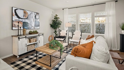 A living room with a black and white checkered rug, white couch with white and burnt orange pillows, a console table under an abstract art piece against the left wall, a wooden coffee table in the middle, and two metal and cloth chairs sitting in front of 3 large windows framed by sheer white curtains.