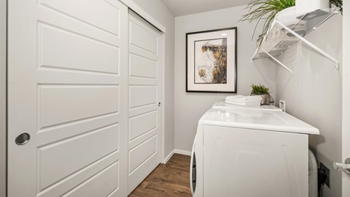 A laundry room with a washer and dryer facing two large closet doors. There is a shelf above the laundry machines holding plants and a picture frame on the far wall.