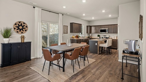 A dining table and kitchen, the dining table is wooden and surrounded by burnt orange chairs that sit on top of a beige rug. The kitchen is in the background and has brown wooden cabinets and a center island.