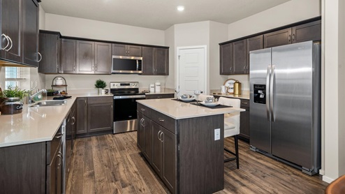 A kitchen with brown wood cabinets, quartz countertops, a refridgerator to the right, a pantry, and an island in the middle.
