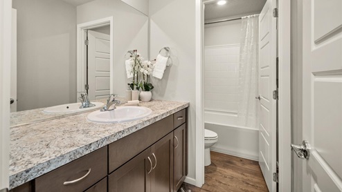 A bathroom with brown wooden cabinets and a patterned countertop. There is a door that opens to a partial view of a toilet and a shower.