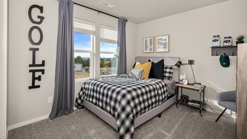 A bedroom featuring a bed with black and white plaid bedsheets and yellow and black pillows. There are picture frames on the wall as well as large black letters spelling the word 'golf'. There are two windows framed by grey curtains.