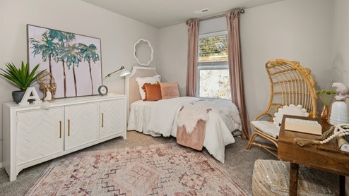 A bedroom with a small bed with white and pink sheets and a large dresser to the left that has a palm tree painting sitting on top. There is a pink ornamental rug and a wicker egg shaped chair in the corner. There is one window framed by pink curtains.