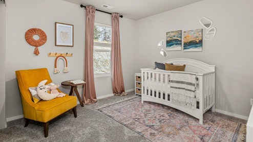 A bedroom with a white crib and a yellow chair that has a moon shaped pillow resting on it. There are paintings, pictures, and wall decor hanging on the walls and one window framed by pink curtains. The crib has a pink ornamental rug underneath.