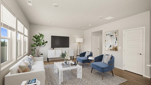 A living area with a grey rug and a white couch with blue accent chairs, a television is hung against the far wall and the left wall has three large windows letting in natural light.