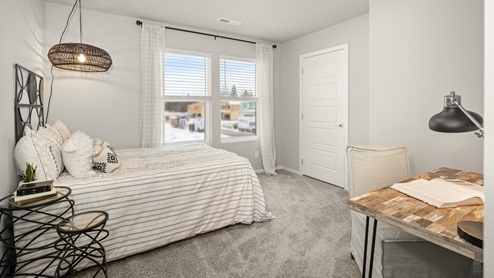 A bedroom featuring a bed with striped white sheets and decorative pillows. There's a nightstand framing the bed and a lamp hanging above the bed. In the right corner is a wooden desk and there are two large windows letting in natural light framed by sheer white curtains.