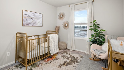 A bedroom featuring a wooden crib and wall art. There's a pink accent chair in the right corner next to a large plant. There's one large window letting in natural light framed by white curtains.