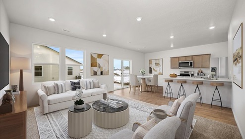 A living area with a large white patterned rug and white couch and grey accent chairs. There's a white coffee table in the middle and a television mounted on the left wall. There's abstract art hanging on the walls and two windows letting in natural light.