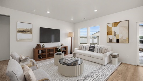 A living area with a large white patterned rug and white couch and grey accent chairs. There's a white coffee table in the middle and a television mounted on the left wall. There's abstract art hanging on the walls and two windows letting in natural light.