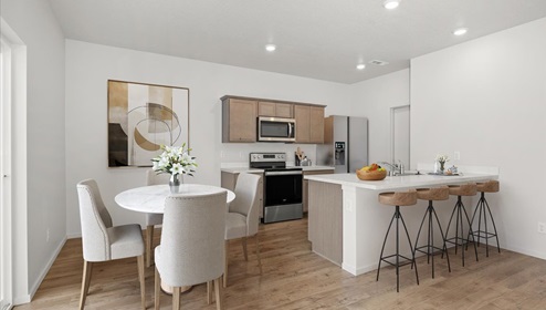A dining area with a round table and white dining chairs and an abstract painting and a kitchen with wood cabinets, stainless steel appliances, and a large peninsula with four wooden stools.