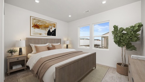 A bedroom with a bed with white and tan sheets in a brown wood bedframe. There are two bedside tables with lamps and abstract art hanging above and two windows letting in natural light with a large plant to the right.