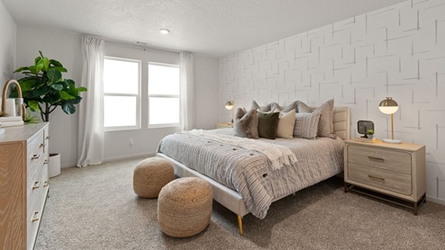 A large bedroom with a bed with grey sheets and decorative pillows, two nightstands with lamps, two ottomans at the foot of the bed, a dresser to the left with a large plant in the corner, and two large windows framed by white curtains.