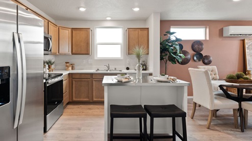 A kitchen with brown wood cabinets, white countertops with an island in the center with two black stools, a fridge to the left, and a dining area to the right.