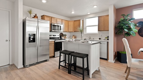A kitchen with brown wood cabinets, white countertops with an island in the center with two black stools, a fridge to the left, and a dining area to the right.