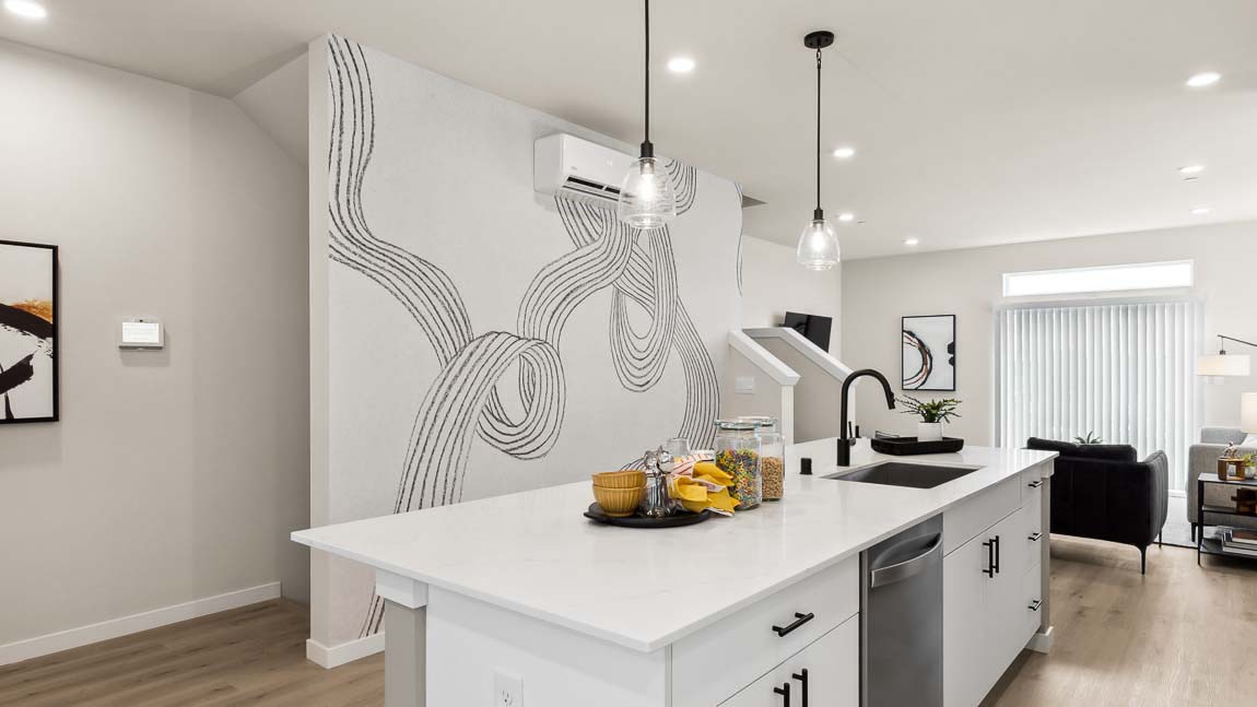 Interior view of model home kitchen with wall artwork, living room in the far background, and showing Smart Home technology screen in stairwell hallway landing
