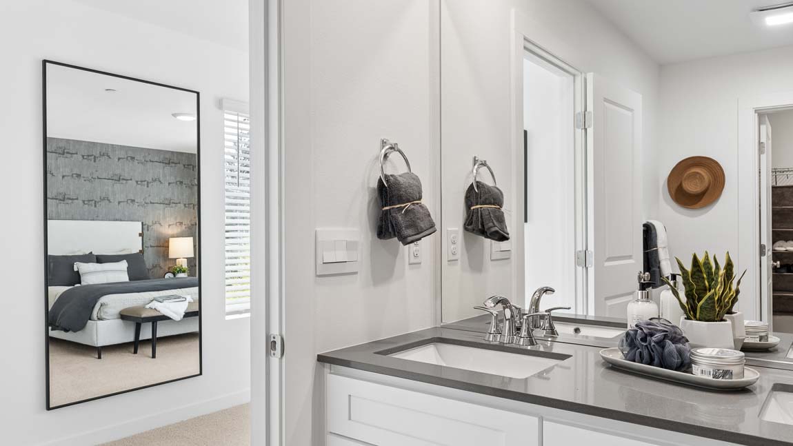 Double-sink bathroom vanity with modern fixtures, large mirror, and view toward the adjacent primary bedroom.
