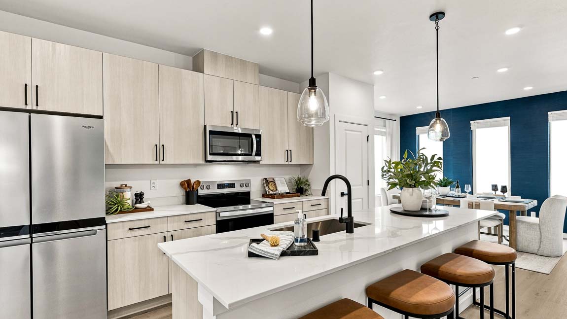 Open-concept kitchen with light wood cabinetry, stainless steel appliances, a white island with brown stools, pendant lights, and a dining area against a dark blue accent wall.