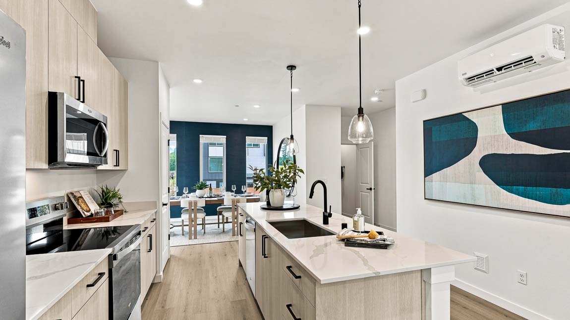 Modern galley-style kitchen with light wood cabinets, stainless steel appliances, a white island with black faucet, pendant lights, and large blue-and-white wall art.