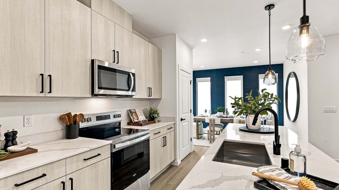 Close view along a modern kitchen island with white counters, a black sink faucet, light wood cabinets, stainless steel appliances, and a dining area with a dark blue wall.
