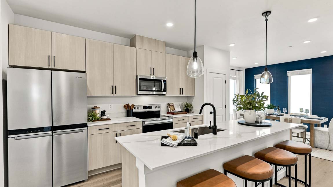 Bright modern kitchen with light wood cabinetry, stainless steel appliances, a white island with brown stools, glass pendant lights, and a dining area in the background.