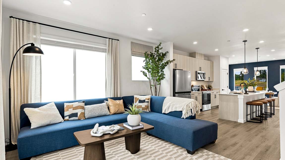 Open-plan living room with a large blue sectional, wood coffee table, patterned rug, tall windows with beige curtains, and the kitchen and island in the background.