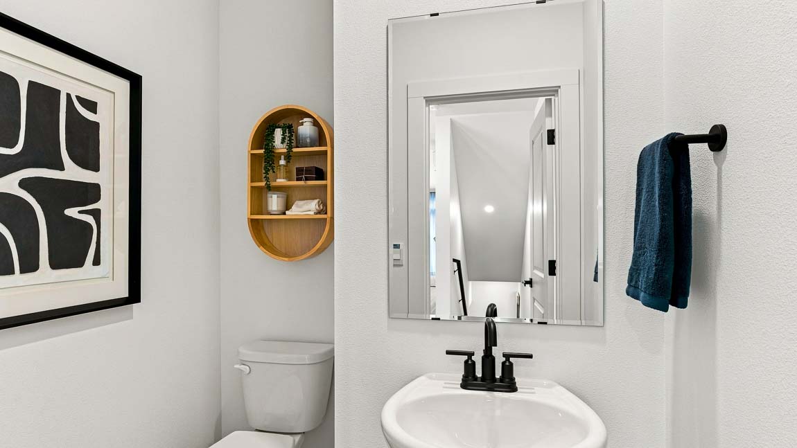 Compact powder room with a white pedestal sink, black faucet, rectangular mirror, toilet, dark blue hand towel, oval wood shelf, and black-and-white framed art.