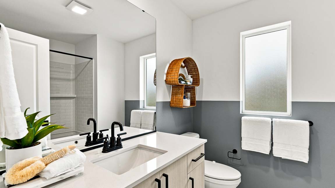 Bathroom with a white vanity and black faucet, large mirror, toilet, frosted window, white towels, woven wall shelf, and a tub-shower with white tile.
