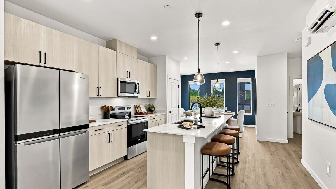 Wide view of a modern kitchen with light wood cabinets, stainless steel appliances, a long white island with four brown stools, and a dining area beyond.