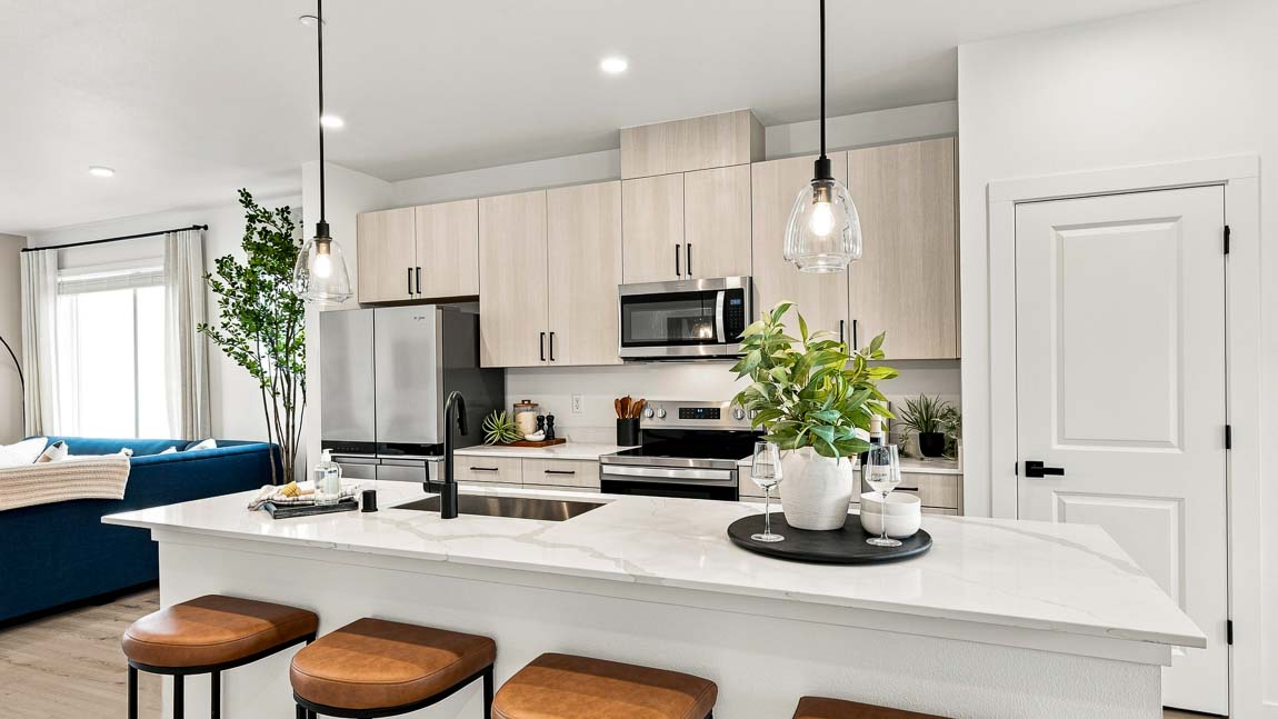 White kitchen island with three brown stools and a potted plant, facing light wood cabinets, stainless steel appliances, pendant lights, and an adjacent blue sofa.