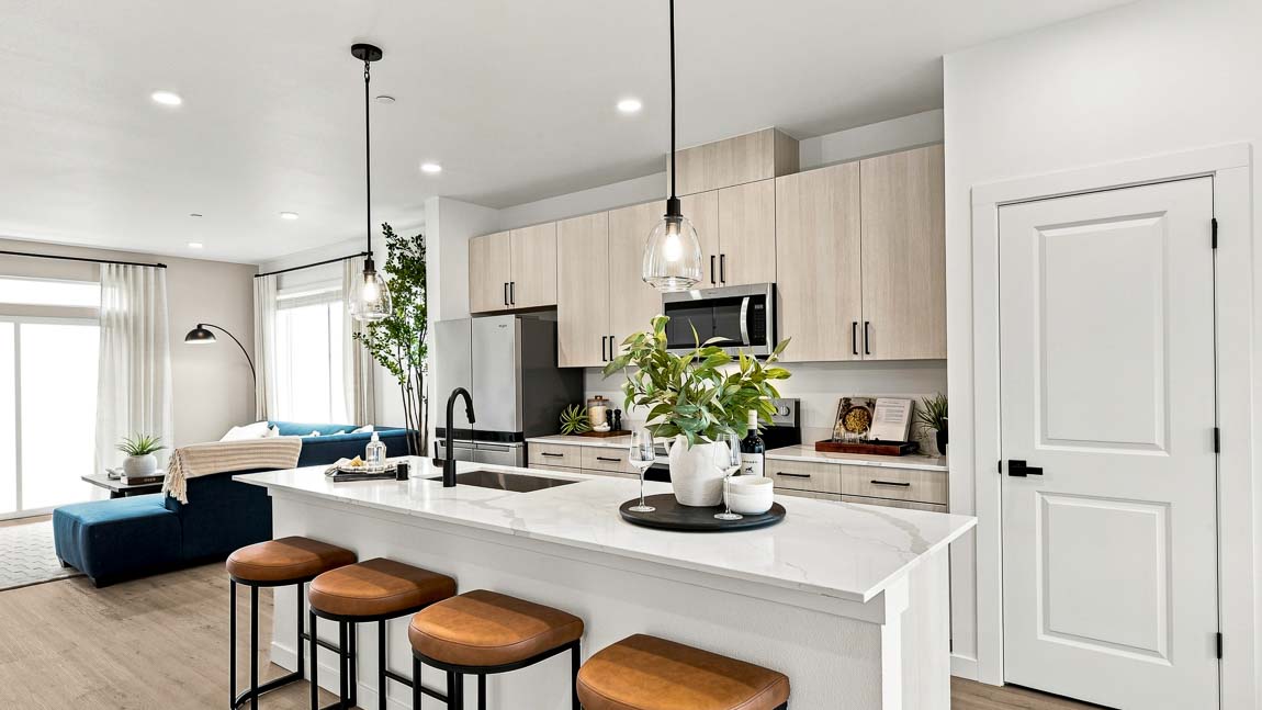 Open kitchen and living area with a white island, four brown stools, potted greenery, light wood cabinets, stainless steel appliances, and a blue sectional sofa.