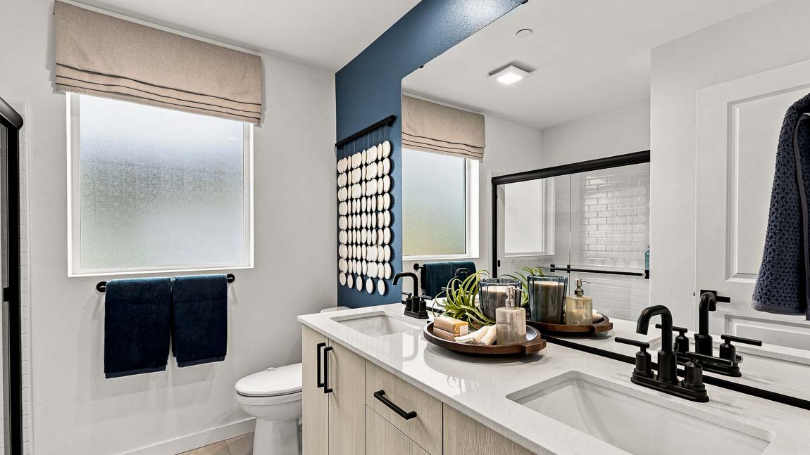 Bathroom with a long double-sink vanity, white countertop, black fixtures, large mirror, blue accent wall, frosted windows, toilet, and glass-enclosed shower.