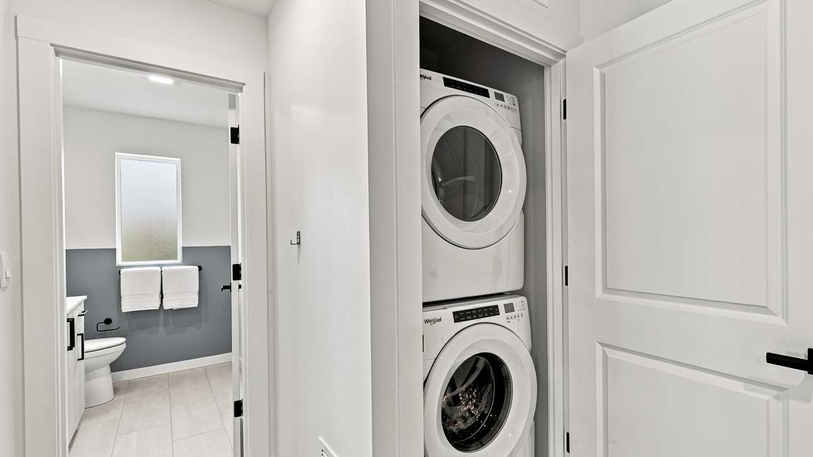 Stacked white washer and dryer in a hallway laundry closet, with an open doorway showing an adjacent bathroom with a toilet, vanity, and frosted window.