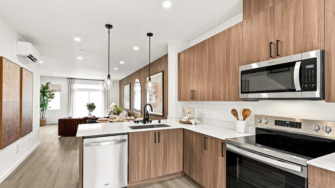 View along a modern kitchen with wood cabinetry, white counters, stainless steel dishwasher, range and microwave, black faucet, pendant lights, and living area beyond.