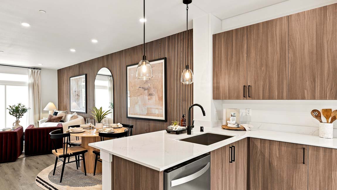 Kitchen island with white quartz countertop, black sink and faucet, pendant lights and dishwasher, opening to a dining table and living area beside a wood slat accent wall.