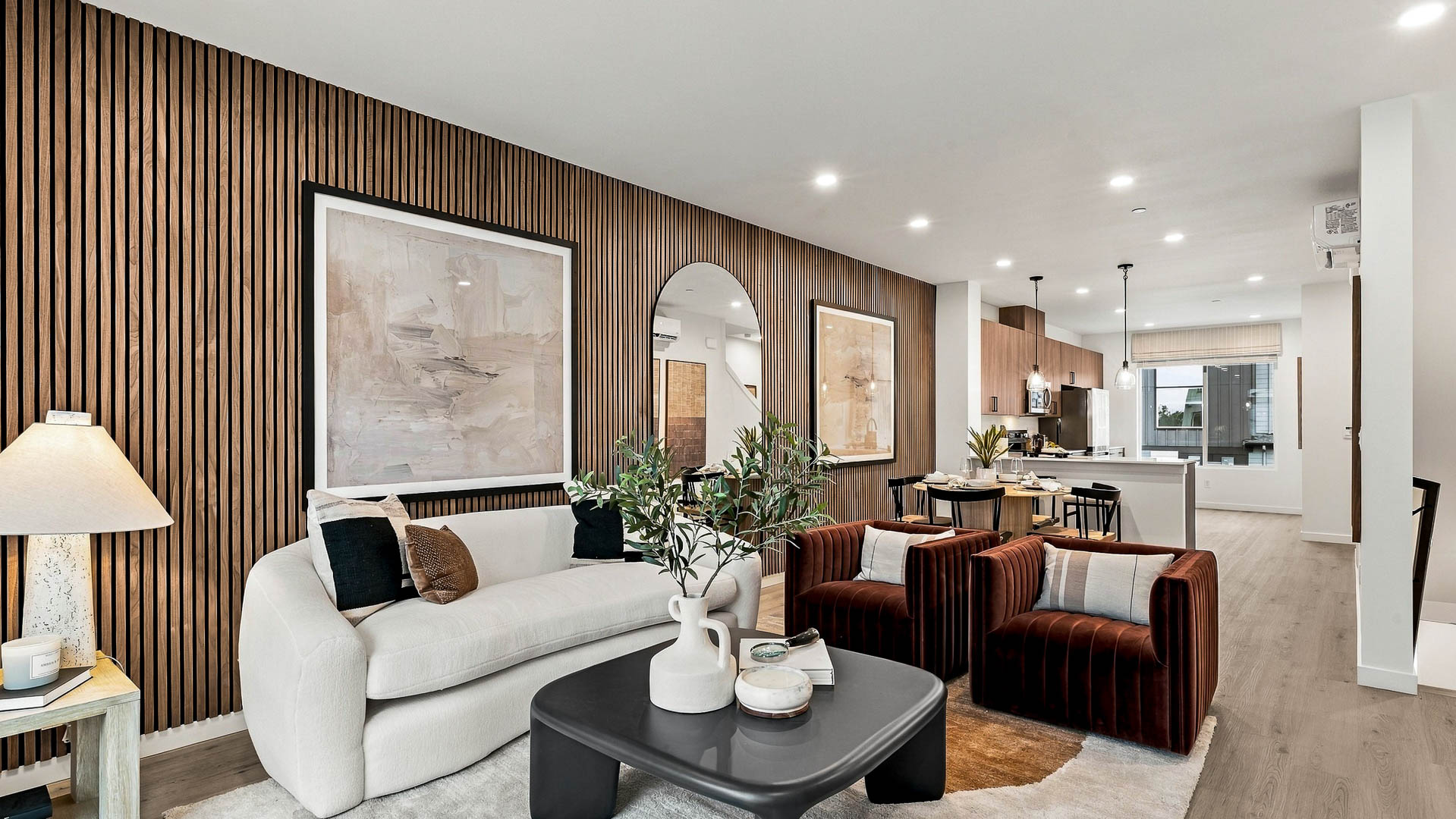 Open-plan living, dining and kitchen space with a white sofa, two rust-colored chairs, black coffee table, wood slat accent wall, round dining table, and kitchen beyond.
