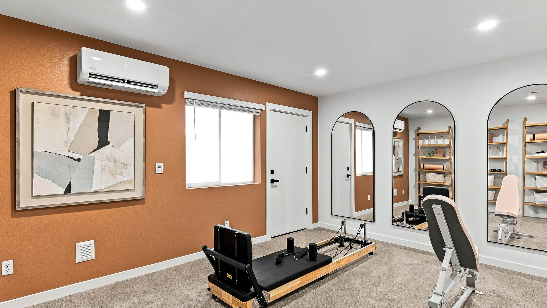 Carpeted home fitness room with a Pilates-style reformer, adjustable exercise bench, three tall arched mirrors, wood shelving, orange accent wall, window, and mini-split unit.